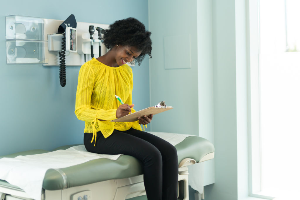 A black woman is at a routine medical appointment. The patient is sitting on a medical examination table in a clinic. She is filling out health history and medical insurance paperwork on a clipboard. The woman is waiting for her doctor.
