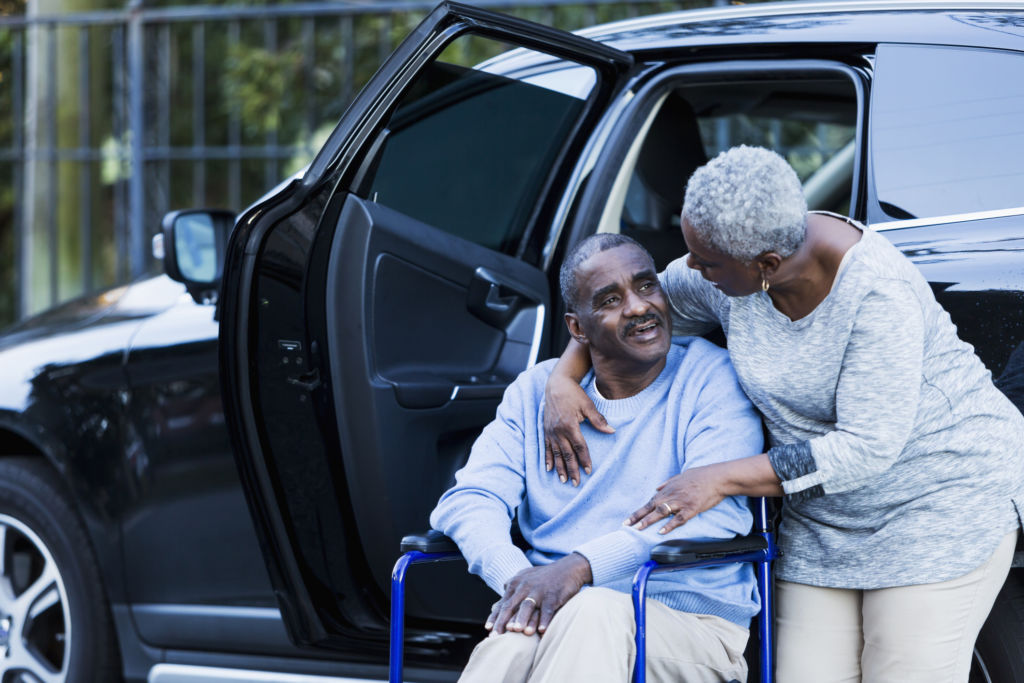 A disabled senior African American man has gotten out of a car, and is sitting in his wheelchair. His loving wife is talking to him with her arm around his neck.