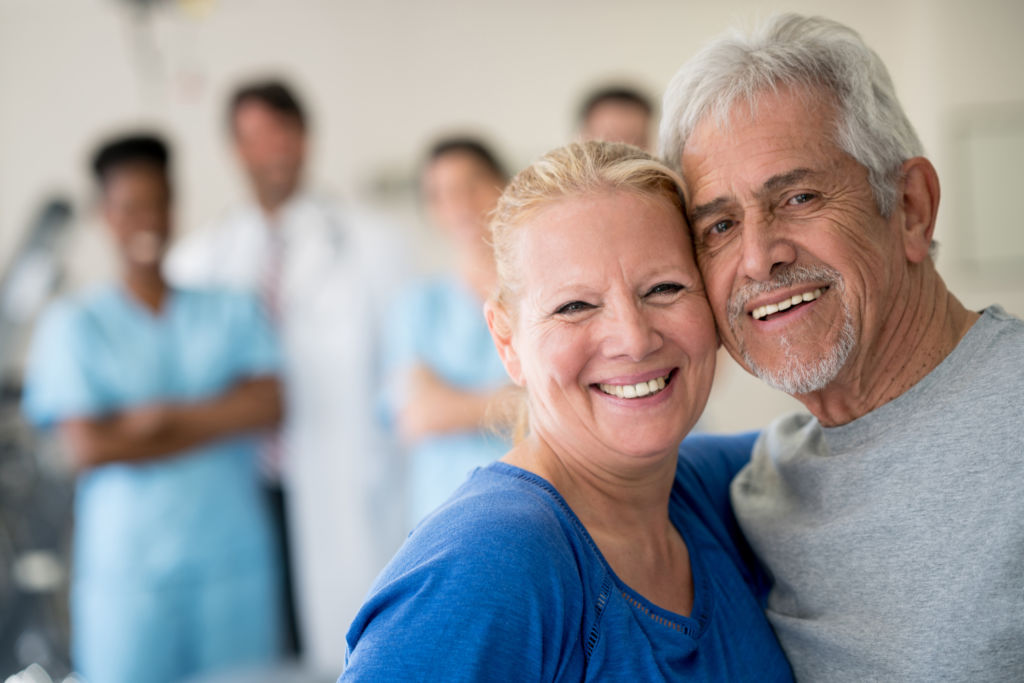 Portrait of senior couple at the clinic leaninig their heads together and looking at the camera smiling while a team of doctors and nurses are standing at the background